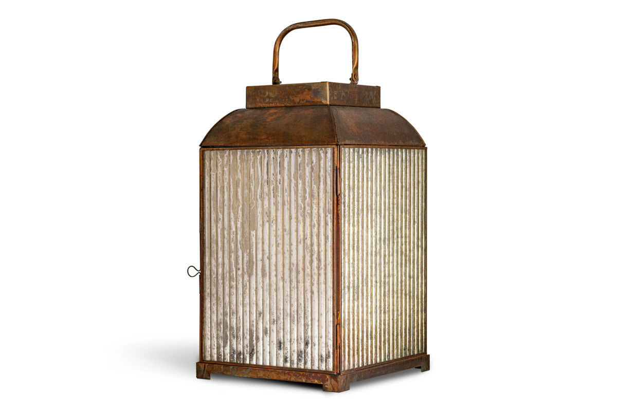 The nkuku Ruala Lantern - Brass & Clear features a vintage-inspired square design with a rusted brass finish, ribbed glass panels, and an aged appearance reminiscent of candlelit evenings. It includes a hinged door with a small latch on one side and a curved handle on top, and is photographed on a flat white background.