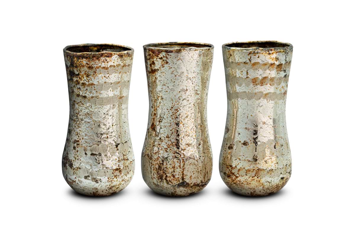 Three Rumi Recycled Glass Mini Vases by nkuku stand side by side against a white background, forming an exquisite decorative set. Each mini vase features a slightly flared rim and a rounded, narrow base, crafted from recycled glass. Their surfaces display a mottled, rustic texture with silvery and brownish hues, showcasing the elegant Silver Rust finish that gives them a vintage, aged look.