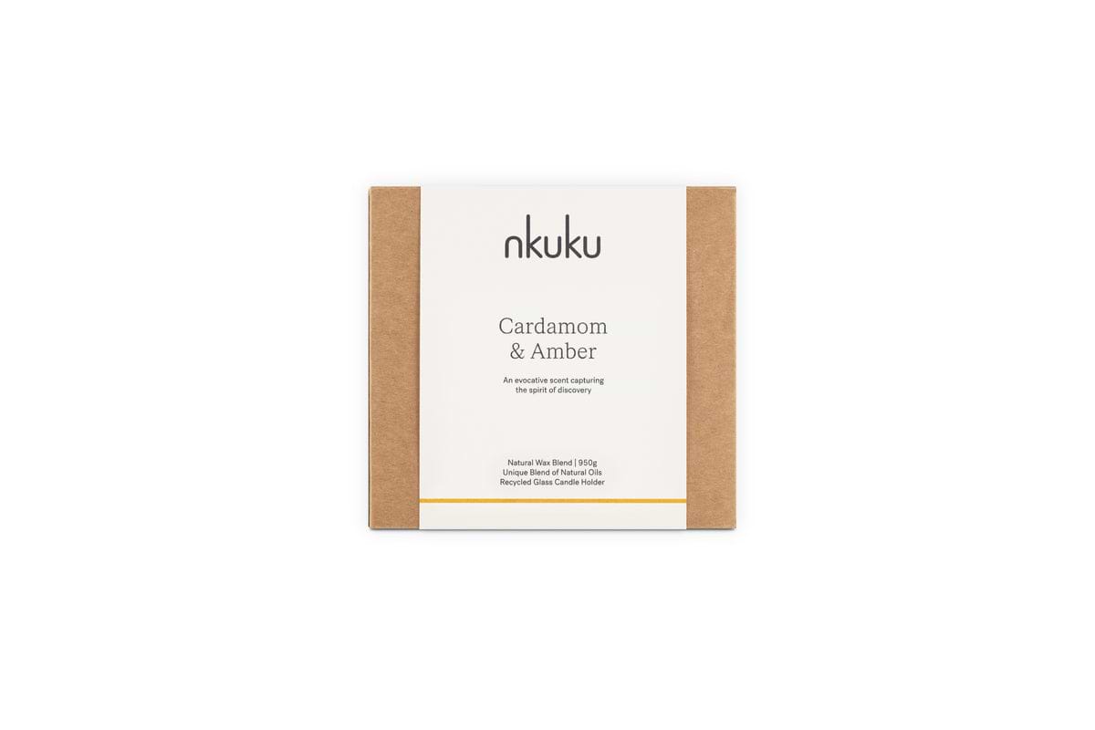 A rectangular box with brown sides and a white label reading “nkuku Cardamom & Amber 3 Wick Scented Candle,” a natural wax blend with sandalwood notes in a recyclable glass holder.