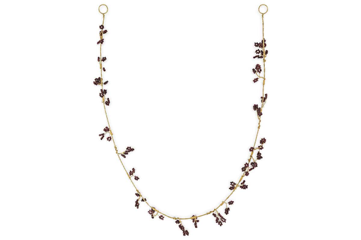 The nkuku Tanu Mango Wood Beaded Garland features brass and ruby beads, artfully clustered along the strand for a whimsical, foliage-inspired look. Displayed in a gentle U shape against a plain white background.