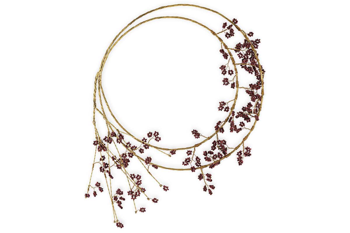 The Tanu Layered Mango Wood Beaded Wreath by nkuku features two intertwined brass-finished branches, adorned with ruby-red beads clustered on the right, while several branch tips extend downward to the left for a festive, minimalist look.
