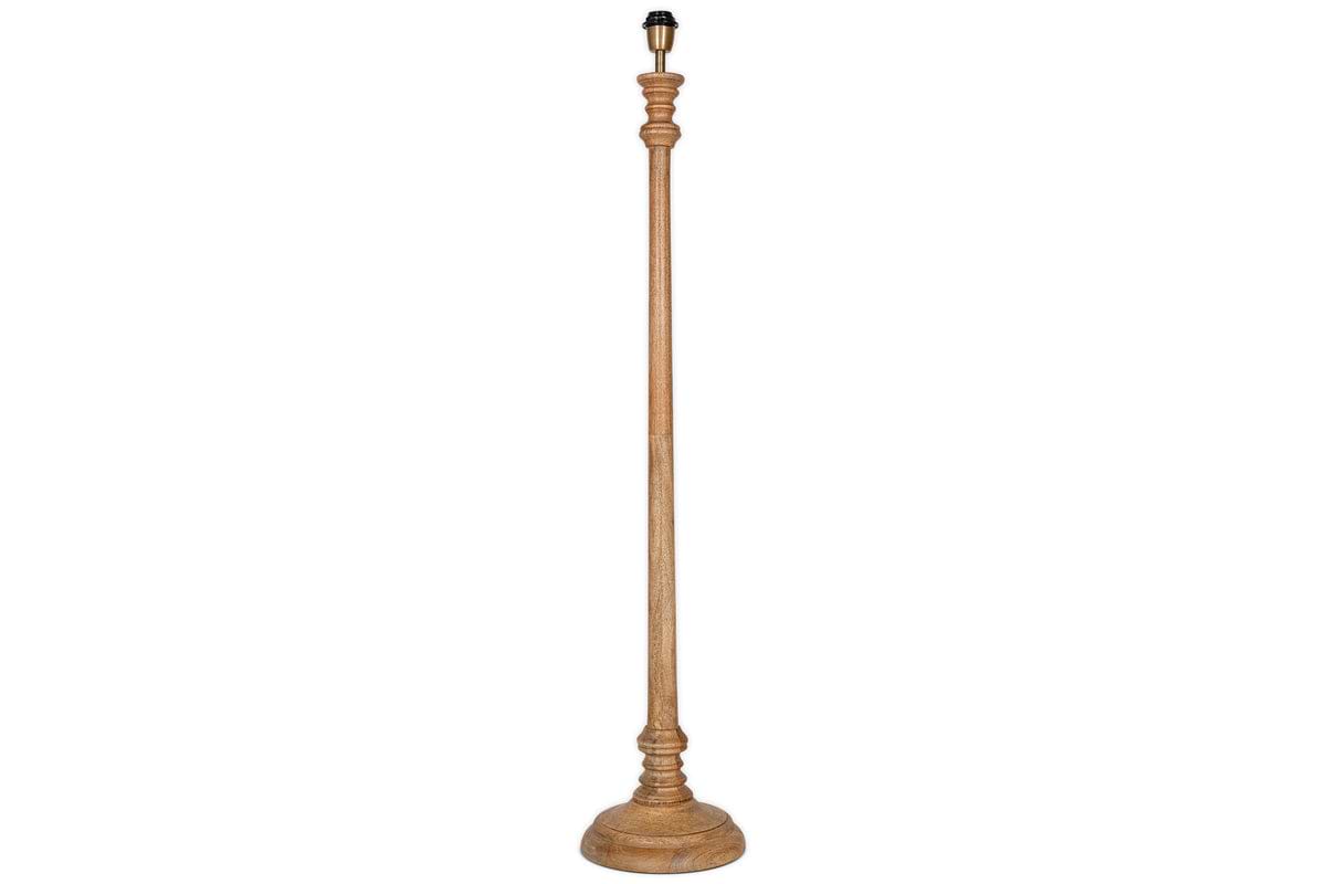 The Usha Classic Wooden Floor Lamp - Natural by nkuku is a tall, handmade mango wood base with a natural finish, round sturdy bottom, and ornately carved slender pole. Eco-friendly, it comes without a lampshade or bulb. Background is white.