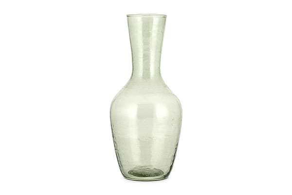 The Yala Hammered Jug - Sage Green by nkuku is a tall, clear glass jug with a narrow neck and wider base, crafted from subtly textured, slightly green-tinted recycled glassware.