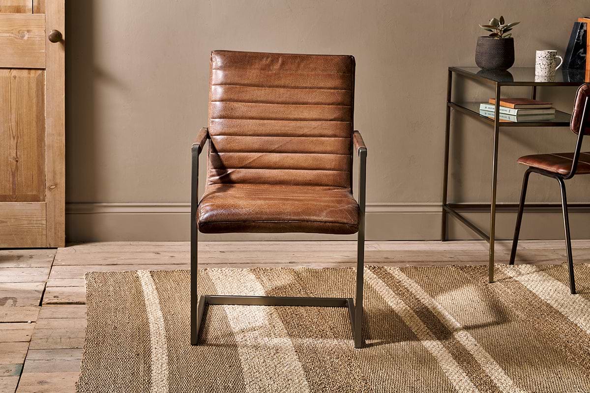 The Wamma Leather Desk Chair by nkuku, featuring distressed dark brown leather and a ribbed design, sits on a jute rug in front of a tan wall.