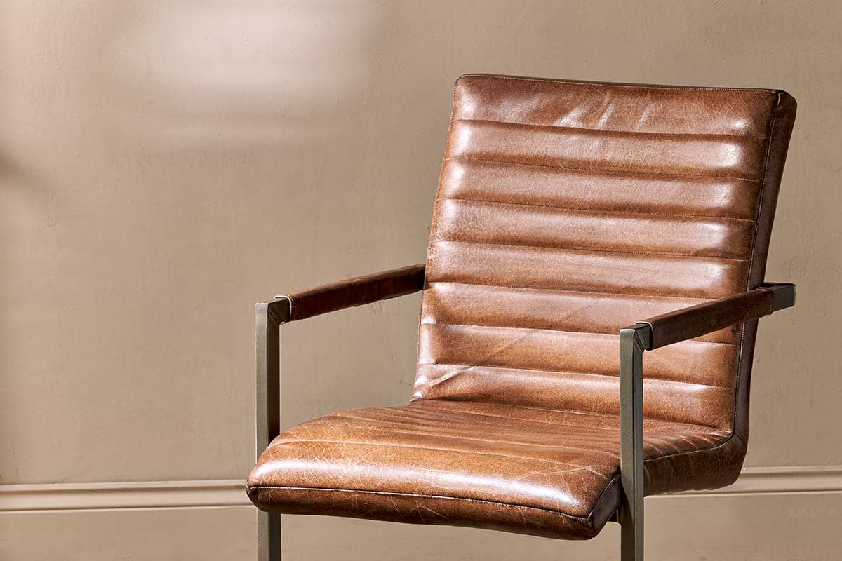 The Wamma Leather Desk Chair by nkuku features dark chocolate brown leather with ribbed design and horizontal channel tufting, slim gunmetal grey metal arms and frame, shown against a warm beige wall.