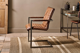 The Wamma Leather Desk Chair by nkuku, featuring quilted dark brown leather and a black metal cantilever frame, sits on a beige striped rug in a room with wooden floors, neutral walls, a wooden door, and a decor-adorned side table.