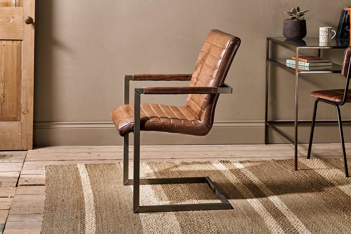 The Wamma Leather Desk Chair by nkuku, featuring quilted dark brown leather and a black metal cantilever frame, sits on a beige striped rug in a room with wooden floors, neutral walls, a wooden door, and a decor-adorned side table.