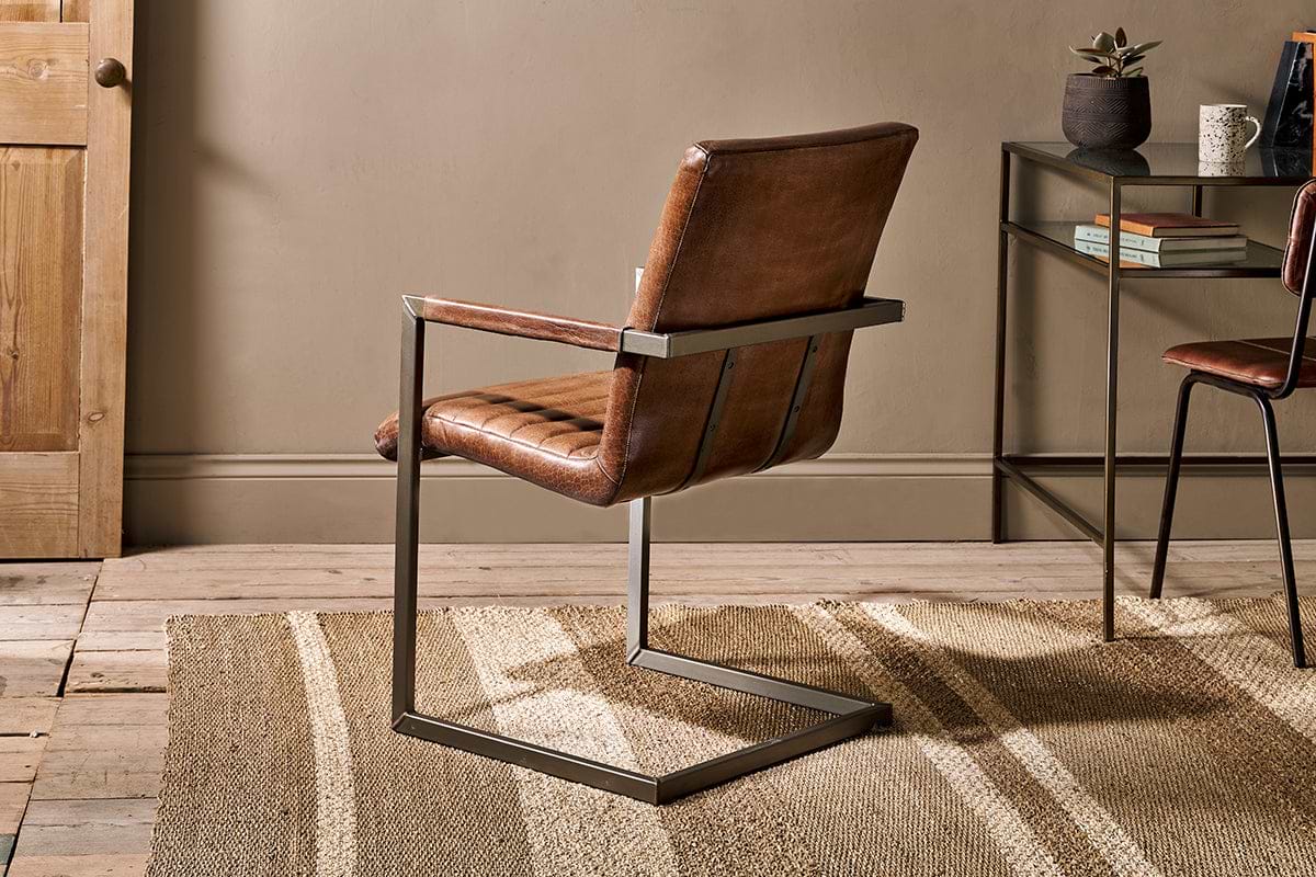 The nkuku Wamma Leather Desk Chair in dark brown with a ribbed design sits on a tan textured rug in a minimal, modern room. Facing away from a light wood door, it’s near a glass table with a plant, mug, and decor by the beige wall.