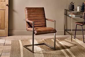 The Wamma Leather Desk Chair by nkuku, featuring dark brown leather with a ribbed design and a cantilever metal frame, sits on a beige textured rug in a cozy room with wood flooring, tan walls, and a glass console table in the background.