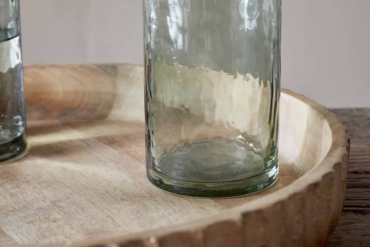 A set of four nkuku Yala Hammered Highball glasses in Sage Green sits on a round, light-finished wooden tray atop a wood surface, with part of another cocktail glass visible to the left.