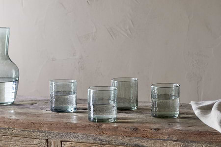 Four Yala Hammered Tumblers in sage green by nkuku, filled with water, rest on a rustic wooden table. A partial glass carafe sits to the left and a white cloth is draped on the right, with a plain textured wall in the background.