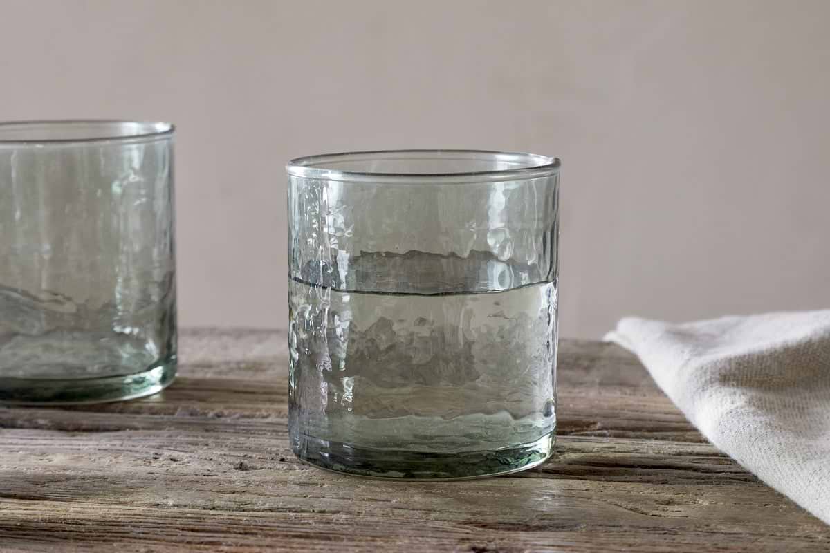 A sage green, hammered recycled glass tumbler from the Yala Hammered Tumbler - Sage Green (Set of 4) by nkuku is placed on a rustic wooden surface, with part of another tumbler and a folded white cloth nearby against a neutral background.