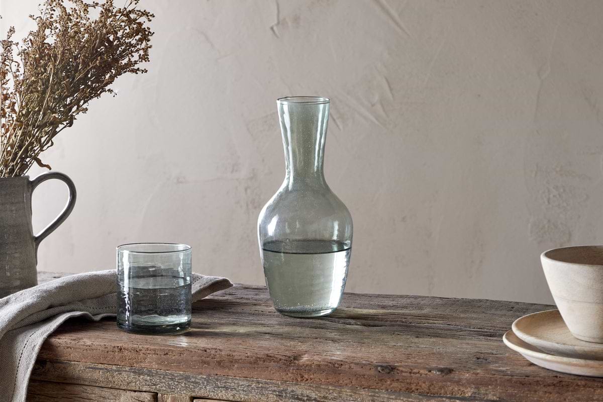 A clear recycled Yala glassware carafe and matching glass, both partially filled with water, sit on a rustic wooden table beside stacked beige bowls and the nkuku Yala Hammered Jug in Sage Green holding dried flowers.
