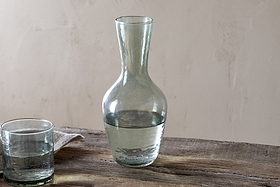 The nkuku Yala Hammered Jug in sage green, partially filled with water, sits on a rustic wooden table beside a glass tumbler and folded cloth, set against a neutral background.