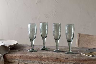 A set of four nkuku Yala Hammered Champagne Glasses in Sage Green is displayed on a rustic wooden table with plates, cutlery, and a napkin nearby, set against a light beige textured wall.