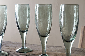 Four Yala Hammered Champagne Glasses in sage green by nkuku stand on a rustic wooden surface against a beige background. The set of four glasses is partially cropped, showing three fully and part of the fourth.