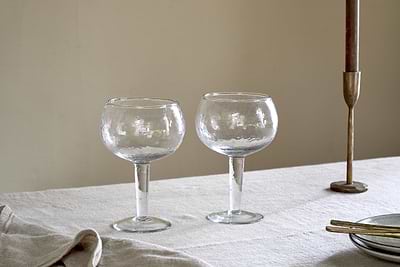 Two nkuku Yala Hammered Gin Glasses (Set of 2) in clear, with textured design, rest on a light tablecloth next to a brass candlestick, gold utensils, and a folded napkin against a plain beige wall.