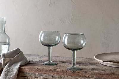 The nkuku Yala Hammered Gin Glass - Sage Green (Set of 2) features two round, clear glasses with green stems on a rustic wooden table, alongside a folded beige cloth, glass carafe, and plate against a textured neutral wall.