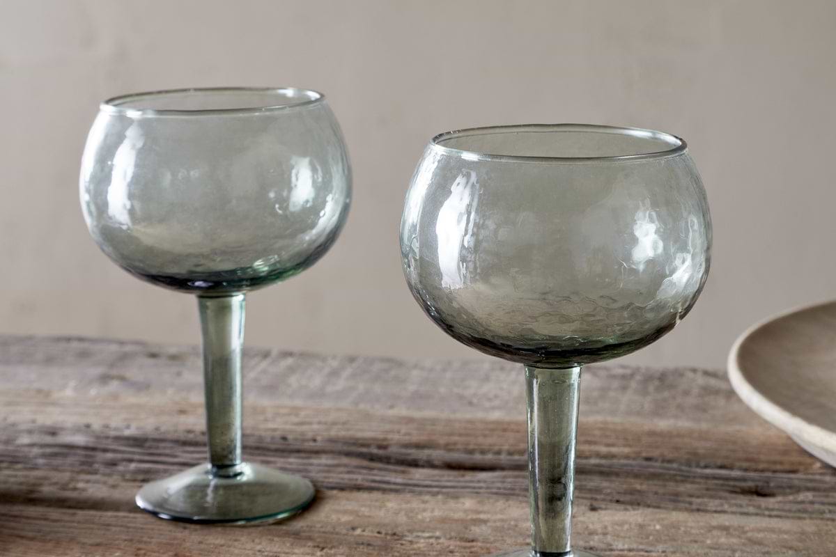 The nkuku Yala Hammered Gin Glasses in sage green, sold as a set of 2, feature round hammered glass and short stems. They sit empty on a rustic wooden surface with a neutral background and a beige plate partially visible.