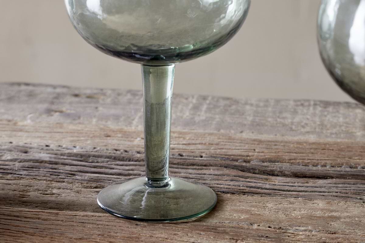 A close-up of the base and stem of an nkuku Yala Hammered Gin Glass in sage green, from a set of 2, displayed on a rustic wooden surface.