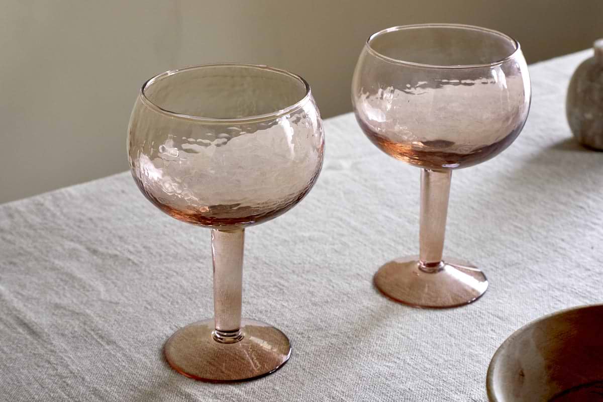 A set of two nkuku Yala Hammered Gin Glasses in Dusty Terracotta rests on a light fabric tablecloth, soft lighting highlighting the textured glass and warm pink tones.