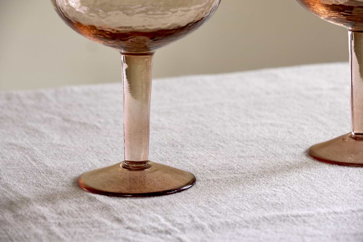 Two Yala Hammered Gin Glasses in Dusty Terracotta by nkuku sit on a light, textured fabric surface, showing only the hammered lower bowls.