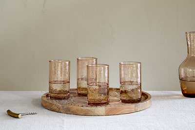 Four nkuku Yala Hammered Highball glasses in Dusty Terracotta (set of 4) sit on a round wooden tray with water, next to a matching carafe and corkscrew, atop a white tablecloth against a light-colored background.