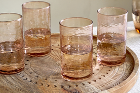 A set of four nkuku Yala Hammered Highball glasses in Dusty Terracotta sits on an ornately carved wooden tray with geometric patterns, displayed on a light-colored surface.