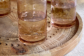 Three nkuku Yala Hammered Highball glasses in Dusty Terracotta, filled with a light brown liquid, sit on a round carved wooden tray with geometric patterns atop a light-colored tablecloth. Set of 4.