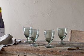 A set of four nkuku Yala Hammered Wine Glasses in Sage Green is arranged on a rustic wooden table with a folded linen napkin, corkscrew, and a partially seen wine bottle against a plain, light-colored wall.
