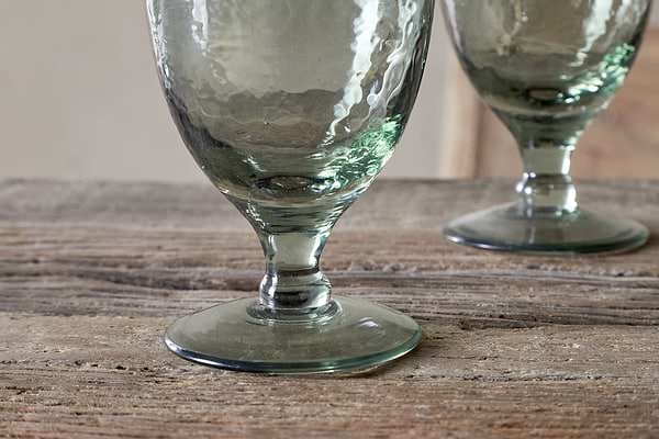Close-up of the nkuku Yala Hammered Wine Glass in Sage Green (Set of 4), featuring thick stems on a rustic wooden surface, with one glass in sharp focus and another softly blurred in the background.