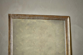A close-up of the Yadur Full Length Mirror by nkuku, featuring an antique brass frame. The mirror is mounted on a light gray textured wall, with only the upper part of the elegant rectangular frame visible.