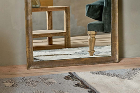 The Yadur Full Length Mirror by nkuku in antique brass reflects a velvet green chair with carved wooden legs and a wooden side table. A textured rug covers the wooden floor, set against neutral-colored walls.