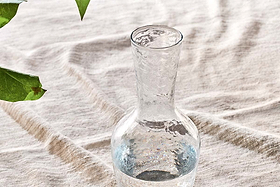 The nkuku Yala Hammered Jug - Clear Glass, filled with water, rests on light wrinkled fabric. A green leafy branch peeks in from the upper left, bringing a natural accent to this calm setting.
