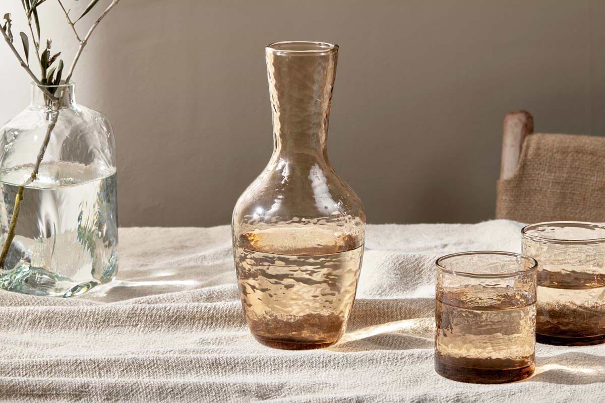 The nkuku Yala Hammered Jug in Dusty Terracotta, filled with water, rests on a beige cloth-covered table with two matching glasses. Nearby, a clear vase with a leafy branch stands next to a wooden chair draped in burlap.