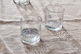The nkuku Yala Hammered Tumblers (Set of 4) feature clear, textured glass and organic reflections as light shines from the left. Shown on a beige, textured fabric tablecloth for a natural aesthetic.