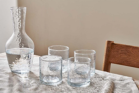 A clear glass carafe with water sits on a textured cloth beside four empty nkuku Yala Hammered Tumblers (Set of 4). A wooden chair appears to the right, against a plain beige wall.