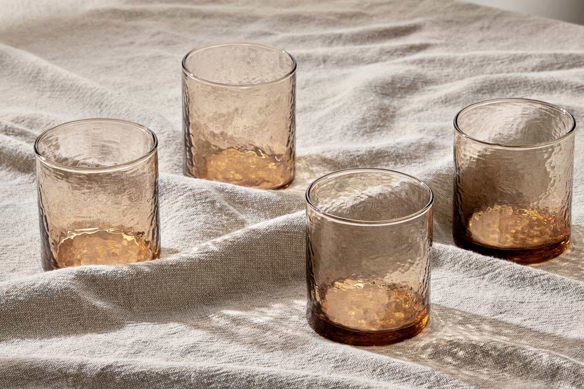 Four Yala Hammered Tumblers in Dusty Terracotta by nkuku, crafted from recycled glass, are arranged on a beige fabric, casting soft shadows and creating a warm, inviting atmosphere. Set includes 4 textured cylindrical glasses.