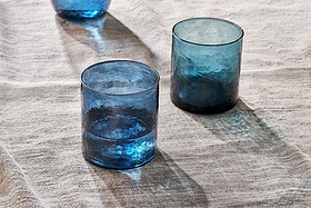 Two nkuku Yala Hammered Tumblers in indigo from the set of 4 rest on a light beige, textured fabric. Sunlight casts soft shadows and highlights, bringing out the glasses’ transparency and the woven look of the surface below.