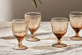 A set of four nkuku Yala Hammered Wine Glasses in Dusty Terracotta sit atop a white, slightly wrinkled tablecloth, sunlight enhancing their translucent, textured glass. A small green leafy stem hangs from the top edge.