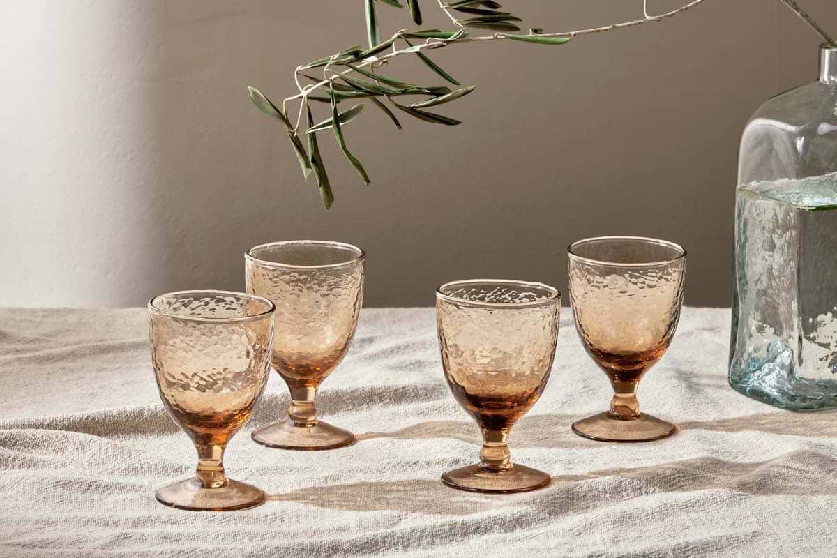Four nkuku Yala Hammered Wine Glasses in Dusty Terracotta are arranged on textured fabric, with an olive branch overhead and a clear vase of water nearby, all illuminated by soft natural light. Set includes 4 glasses.