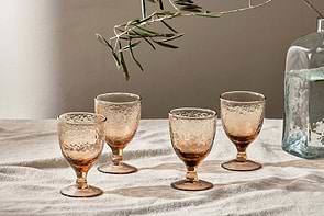 Four nkuku Yala Hammered Wine Glasses in Dusty Terracotta are arranged on textured fabric, with an olive branch overhead and a clear vase of water nearby, all illuminated by soft natural light. Set includes 4 glasses.