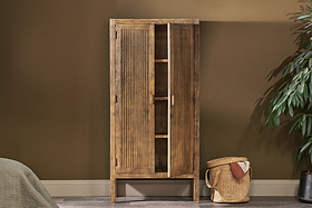 The nkuku Yasha Reeded Mango Wood Cabinet stands against a brown wall, one door slightly open to reveal shelves. A leafy green plant sits to the right, a bed edge is on the left, and a woven basket with a towel rests in front.