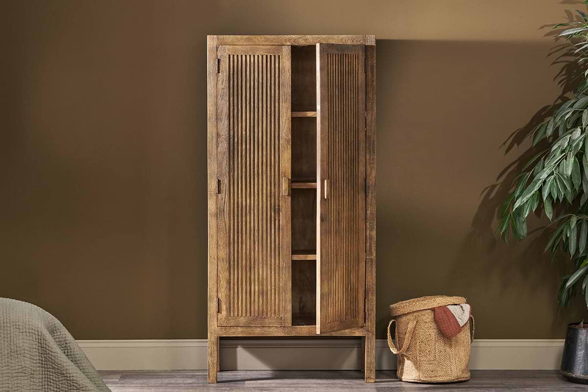 The nkuku Yasha Reeded Mango Wood Cabinet stands against a brown wall, one door slightly open to reveal shelves. A leafy green plant sits to the right, a bed edge is on the left, and a woven basket with a towel rests in front.