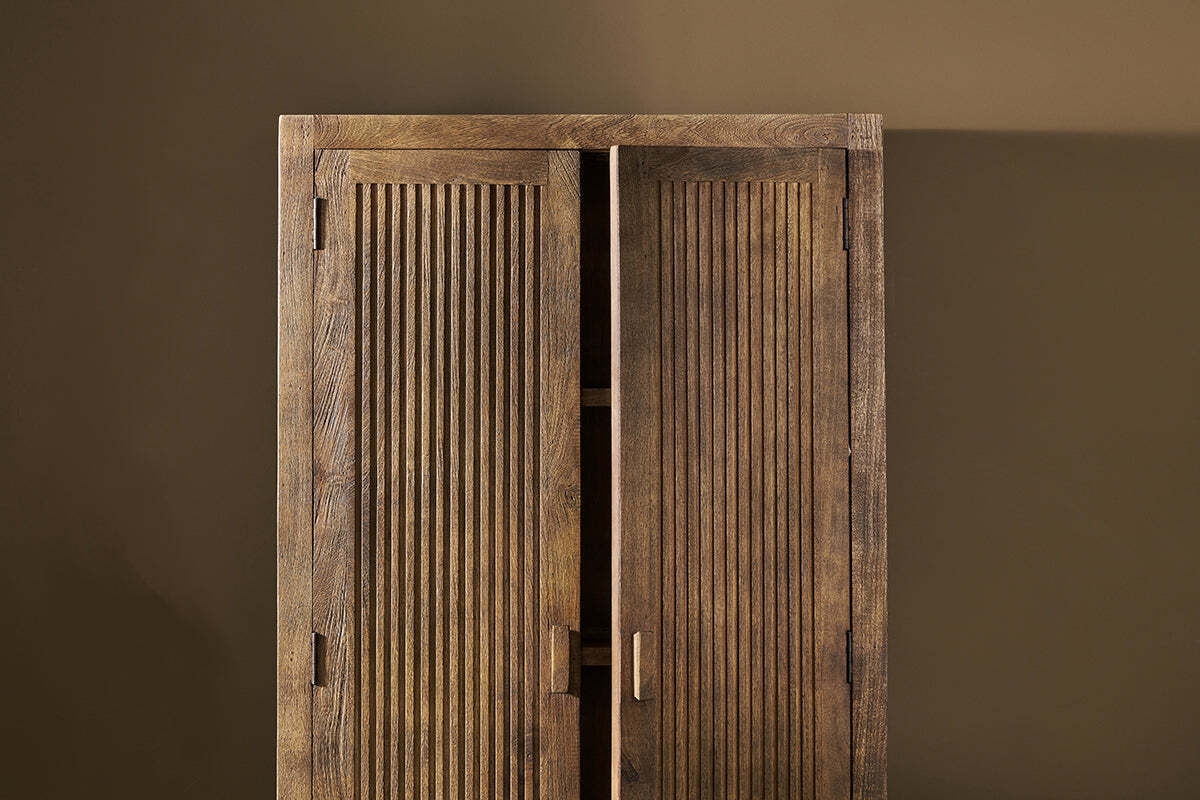 The Yasha Reeded Mango Wood Cabinet by nkuku stands against a plain brown wall. One grooved door is partially open, showing the dark interior. It has a natural textured finish and simple rectangular handles for understated elegance.
