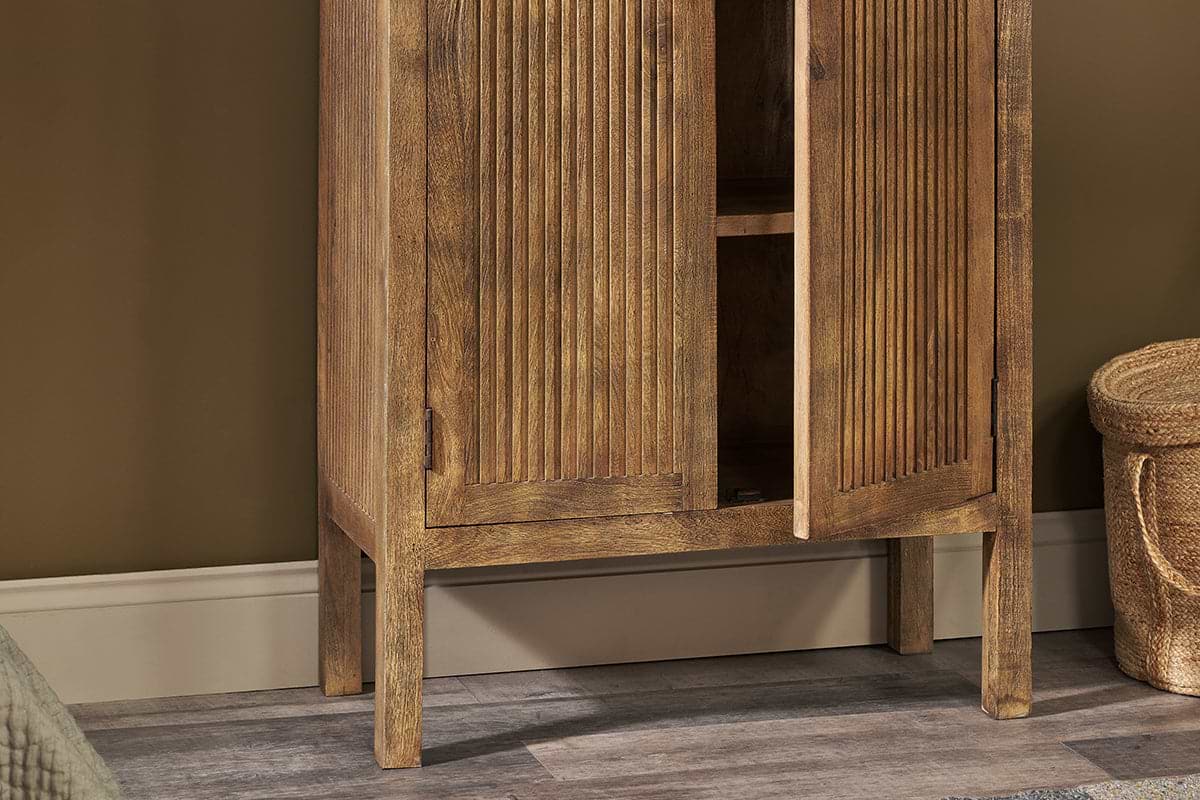 A close-up of the nkuku Yasha Reeded Mango Wood Cabinet with slatted doors, one ajar to reveal shelves. The cabinet stands on four legs over a wood-patterned floor, beside a beige wall and a woven basket partly in view.