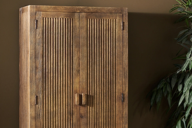 The nkuku Yasha Reeded Mango Wood Cabinet, featuring vertical grooves on two doors, square handles, and visible wood grain, stands against a brown wall—ideal for small spaces. A leafy green plant appears on the right side of the image.