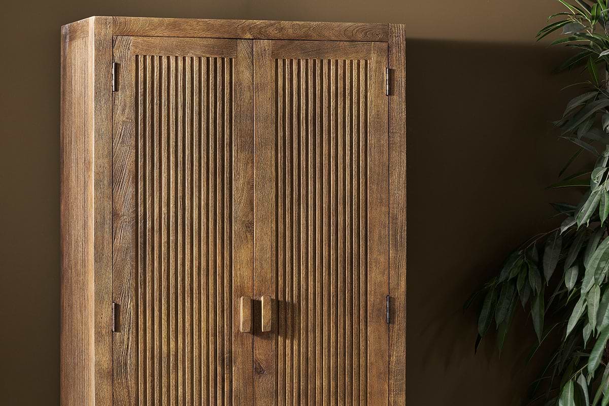 The nkuku Yasha Reeded Mango Wood Cabinet, featuring vertical grooves on two doors, square handles, and visible wood grain, stands against a brown wall—ideal for small spaces. A leafy green plant appears on the right side of the image.