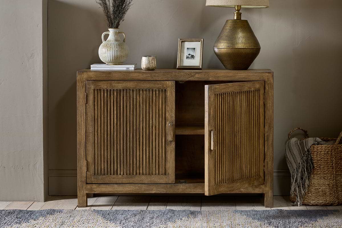 The nkuku Yasha Reeded Mango Wood Sideboard with ribbed doors, one ajar, stands by a beige wall. On top: lamp, framed photo, vase of dried flowers, candle, and book. A woven basket is beside it on a wooden floor with a patterned rug.
