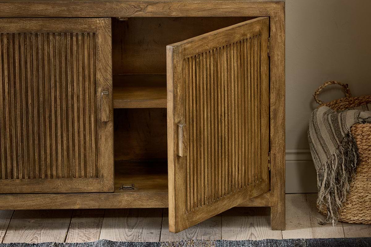 The nkuku Yasha Reeded Mango Wood Sideboard features fluted doors, one slightly open to reveal shelves inside. To the right sits a woven basket with a striped throw.